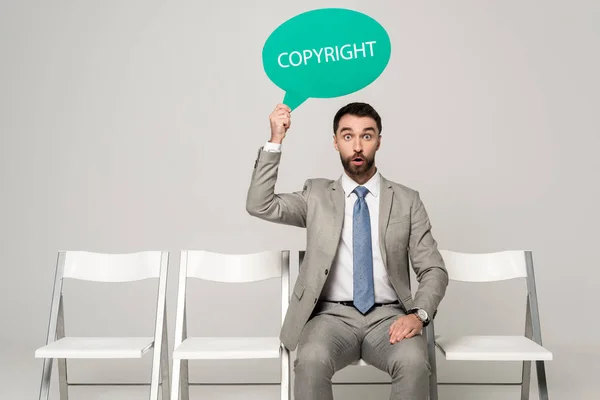 Surprised businessman holding thought bubble with word copyright while sitting on chair on grey background — Stock Photo