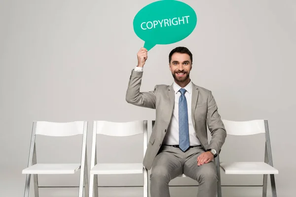 Sonriente hombre de negocios sosteniendo la burbuja del pensamiento con derechos de autor palabra mientras está sentado en la silla sobre fondo gris - foto de stock