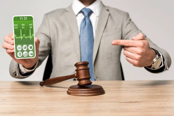 Cropped view of lawyer pointing with finger at smartphone with heartbeat rate app, and gavel on desk — Stock Photo