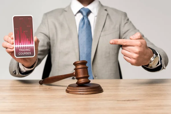 Cropped view of lawyer pointing with finger at smartphone with trading courses app, and judge gavel on desk — Stock Photo