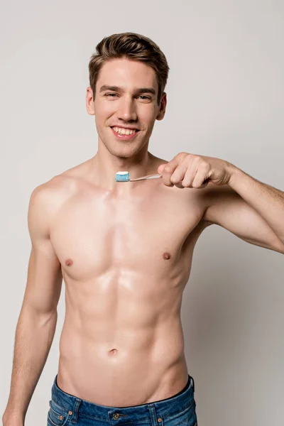 Sonriente hombre sexy con torso desnudo sosteniendo cepillo de dientes aislado en gris - foto de stock