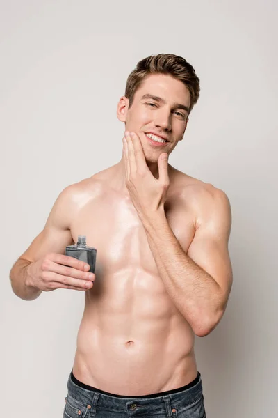 Sonriente hombre sexy con torso muscular aplicando aftershave en la cara aislado en gris - foto de stock