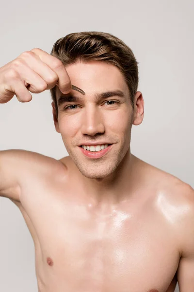 Улыбающийся сексуальный мужчина с голым торсом, щипающий брови пинцетом, изолированным на сером — стоковое фото