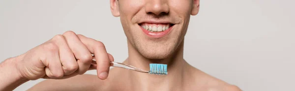 Обрезанный вид улыбающегося сексуального мужчины с обнаженным туловищем, держащего зубную щетку изолированным на сером, панорамный снимок — стоковое фото