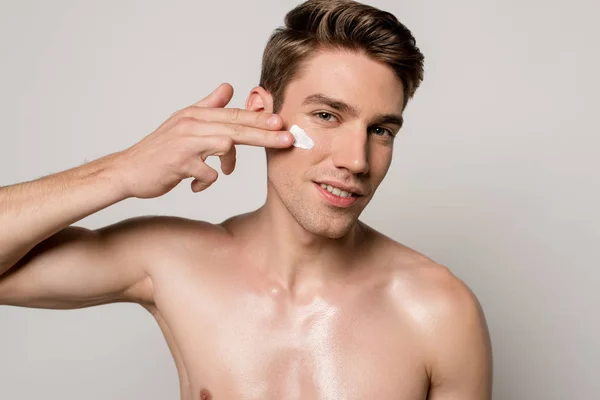 Sonriente sexy hombre con torso muscular aplicando crema facial aislado en gris - foto de stock