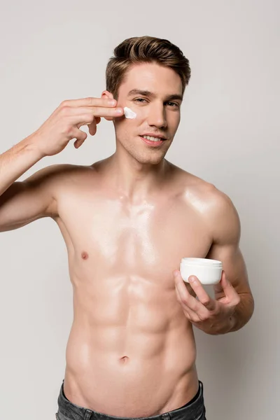 Sorrindo homem sexy com torso muscular aplicando creme facial isolado no cinza — Fotografia de Stock