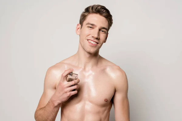 Smiling sexy man with muscular torso spraying perfume isolated on grey — Stock Photo