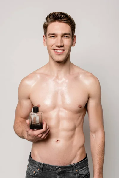 Smiling sexy man with muscular torso holding perfume bottle isolated on grey — Stock Photo