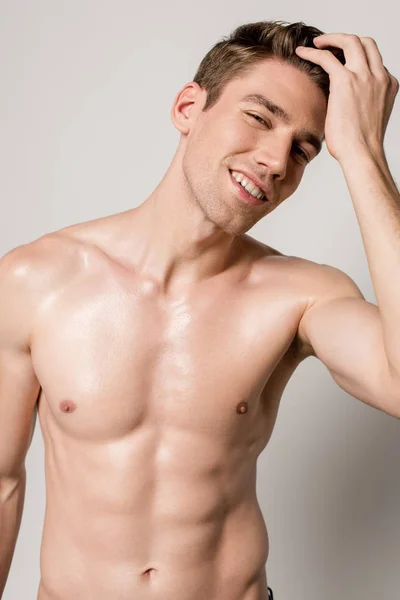 Sonriente sexy hombre con torso muscular tocando el pelo aislado en gris - foto de stock