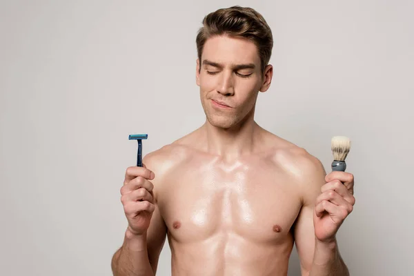 Вдумчивый сексуальный мужчина с мускулистым туловищем проведение бритвы и кисти для бритья изолированы на сером — стоковое фото