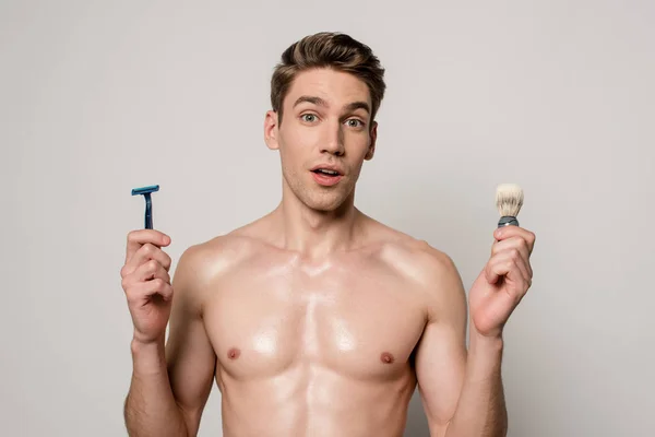 Shocked sexy man with muscular torso holding shaver and shaving brush isolated on grey — Stock Photo