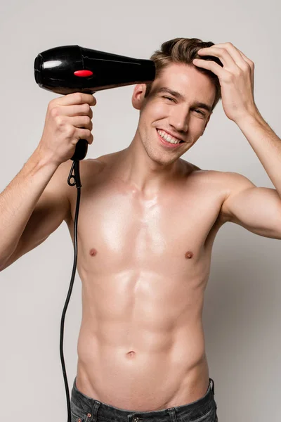Uomo sexy sorridente con busto muscoloso asciugatura capelli con asciugacapelli isolato su grigio — Foto stock