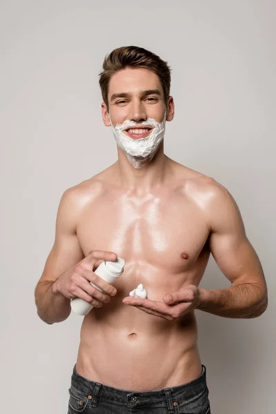 Sorrindo homem sexy com torso muscular aplicando espuma de barbear em mãos isoladas em cinza — Fotografia de Stock