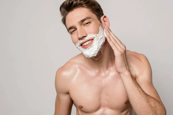 Улыбающийся сексуальный мужчина с мускулистым туловищем нанося пену для бритья изолированную на серый — стоковое фото