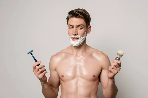 Uomo sexy con busto muscolare e schiuma da barba sul viso tenendo rasoio e spazzola da barba isolato su grigio — Foto stock