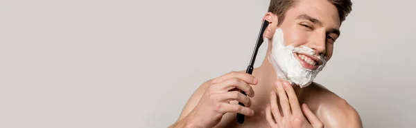 Smiling sexy man with muscular torso shaving with straight razor isolated on grey, panoramic shot — Stock Photo