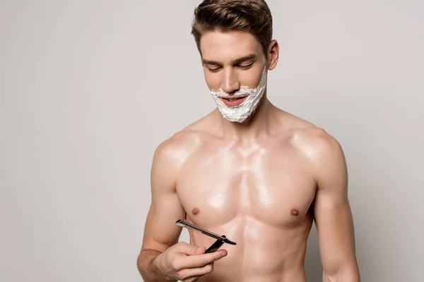 Smiling sexy man with muscular torso and shaving foam on face holding straight razor isolated on grey — Stock Photo