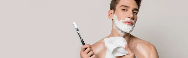 Sexy man with muscular torso and shaving foam on face holding straight razor and towel isolated on grey, panoramic shot — Stock Photo