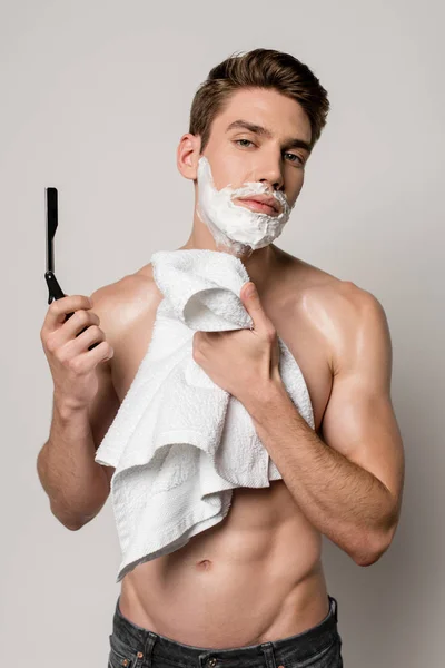 Sexy hombre con torso muscular y espuma de afeitar en la cara celebración de afeitadora recta y toalla aislada en gris - foto de stock