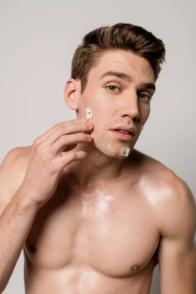 Sexy man with muscular torso touching bloody wounds after shaving on face isolated on grey — Stock Photo