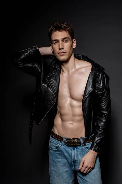 Sexy young man with muscular torso in biker jacket and jeans on black background — Stock Photo