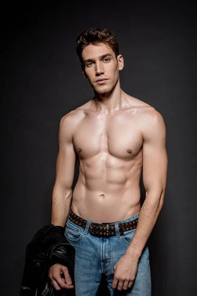 Сексуальный молодой человек с мускулистым туловищем в джинсах, держащий байкерскую куртку на черном фоне — стоковое фото