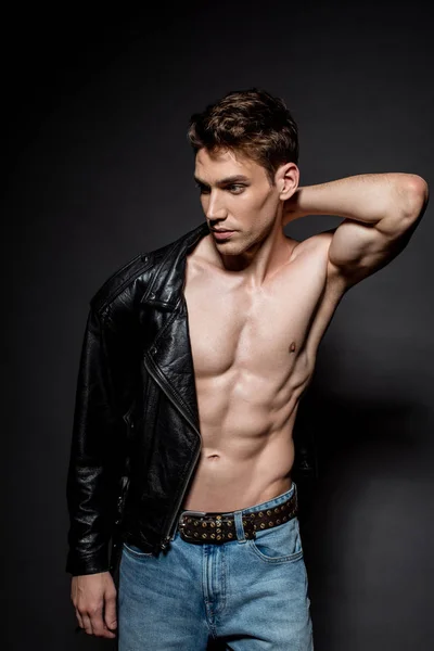 Сексуальный молодой человек с мускулистым туловищем надевает байкерскую куртку и смотрит в сторону на черном фоне — стоковое фото