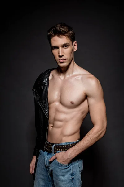 Sexy young man with muscular torso in biker jacket and jeans standing with hand in pocket on black background — Stock Photo