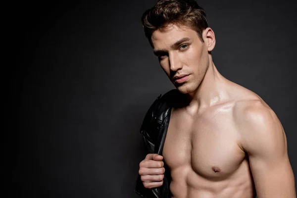 Sexy young man with muscular torso and biker jacket on shoulder on black background — Stock Photo