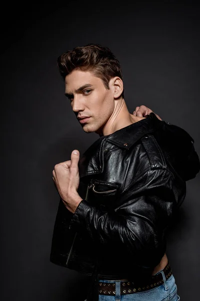 Sexy young man with muscular torso in biker jacket looking at camera on black background — Stock Photo
