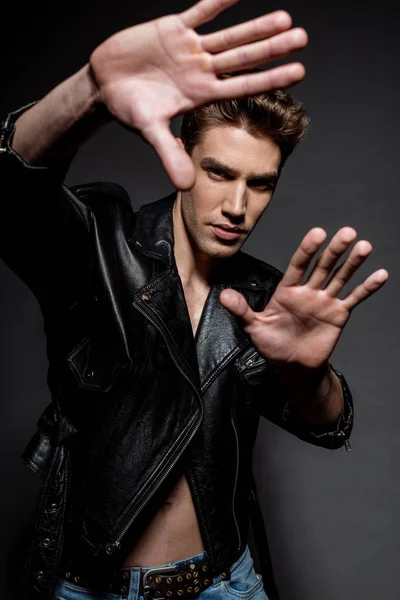 Selective focus of sexy young man with muscular torso in biker jacket and jeans gesturing with hands on black background — Stock Photo
