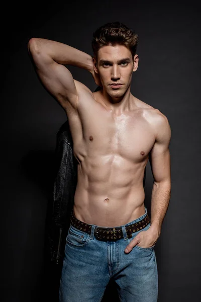 Sexy junger Mann mit muskulösem Oberkörper und Bikerjacke posiert mit der Hand in der Tasche auf schwarzem Hintergrund — Stockfoto