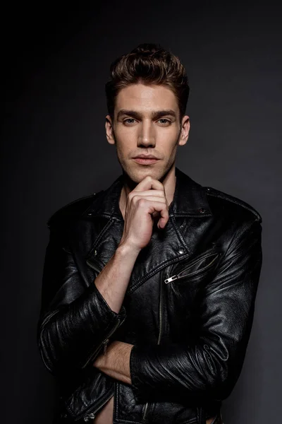 Красивый сексуальный молодой человек в байкерской куртке трогает подбородок на черном фоне — стоковое фото
