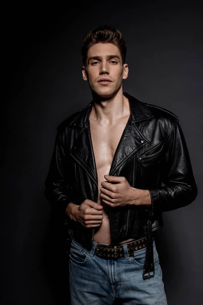 Sexy young man with muscular torso in biker jacket and jeans on black background — Stock Photo