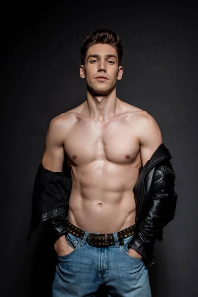 Sexy young man with muscular torso in biker jacket and jeans posing with hands in pockets on black background — Stock Photo