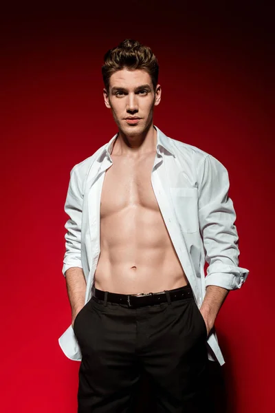 Sexy joven elegante hombre en camisa desabotonada con torso desnudo muscular y las manos en bolsillos sobre fondo rojo — Stock Photo