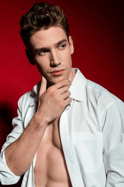 Сексуальный молодой элегантный мужчина в расстегнутой рубашке с мускулистым туловищем касающимся подбородка и отводящим взгляд на красный фон — стоковое фото
