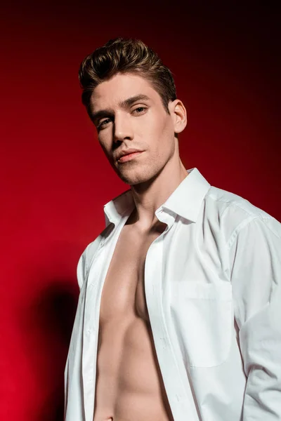 Sexy young elegant man in unbuttoned shirt with muscular bare torso on red background — Stock Photo