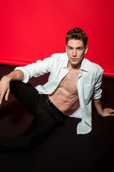 Sexy young elegant man in unbuttoned shirt with muscular bare torso sitting on floor — Stock Photo