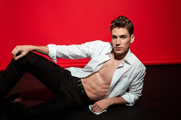 Sexy young elegant man in unbuttoned shirt with muscular bare torso lying on floor on red background — Stock Photo