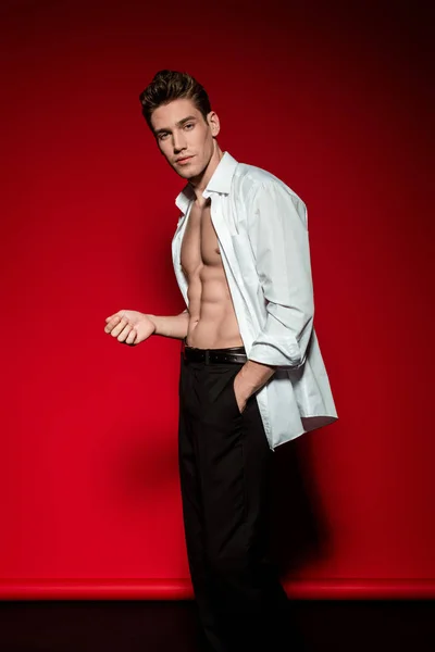 Sexy jovem elegante homem em camisa desabotoada com muscular tronco nu posando com a mão no bolso no fundo vermelho — Fotografia de Stock