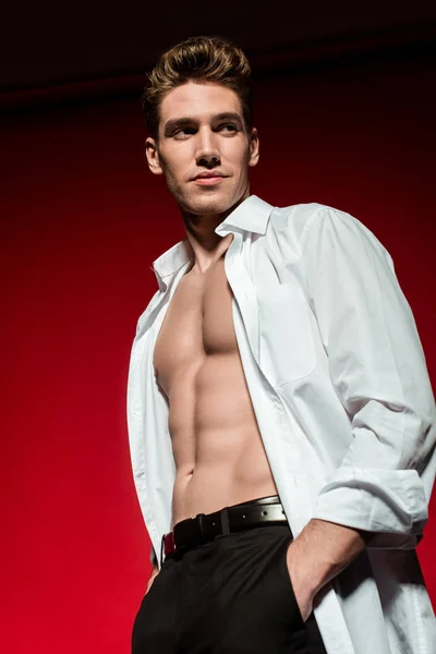 Low angle view of sexy young elegant man in unbuttoned shirt with muscular bare torso posing with hands in pockets on red background — Stock Photo