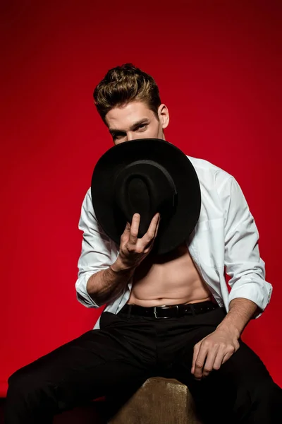 Sexy joven elegante hombre en camisa desabotonada con torso desnudo muscular posando con sombrero cerca de la cara sobre fondo rojo - foto de stock