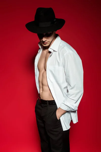 Sexy young elegant man in unbuttoned shirt and hat with muscular bare torso posing on red background — Stock Photo