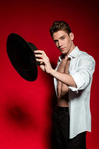 Sexy joven elegante hombre en camisa desabotonada con torso desnudo muscular sosteniendo sombrero sobre fondo rojo - foto de stock