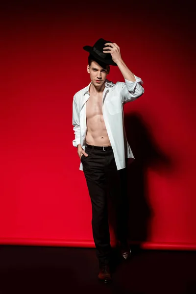 Sexy young elegant man in unbuttoned shirt with muscular bare torso putting on hat on red background — Stock Photo