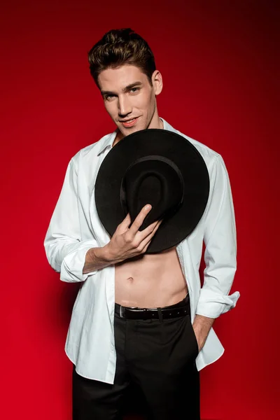 Sexy giovane uomo elegante in camicia sbottonata con busto nudo muscolare e mano in tasca tenendo il cappello su sfondo rosso — Foto stock