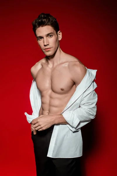 Sexy joven elegante hombre en camisa desabotonada con torso desnudo muscular sobre fondo rojo - foto de stock