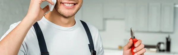 Panoramic shot of happy installer talking on smartphone and holding pliers — Stock Photo