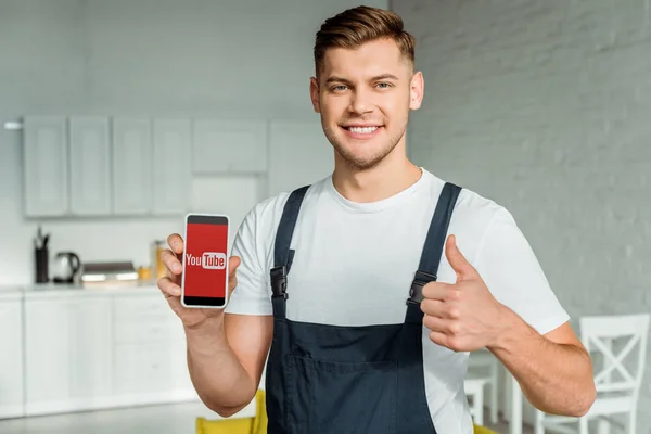 KYIV, UCRANIA - 4 de diciembre de 2019: instalador feliz sosteniendo el teléfono inteligente con la aplicación de youtube en la pantalla y mostrando el pulgar hacia arriba - foto de stock